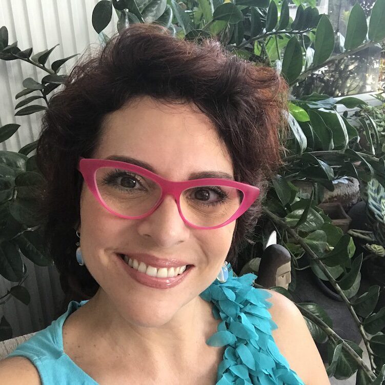 Smiling woman with pink glasses in a lush green setting.