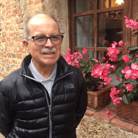 An elderly man smiling near vibrant pink flowers outdoors.