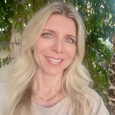 Smiling woman with long blonde hair outdoors under leafy trees.