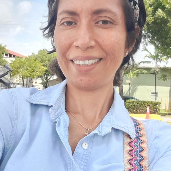 Smiling woman outdoors with trees and buildings in the background.