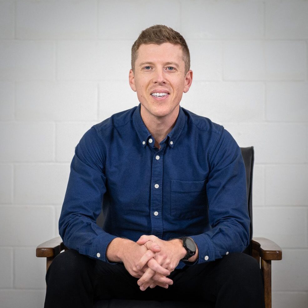 Smiling man in a blue shirt sitting with hands clasped.